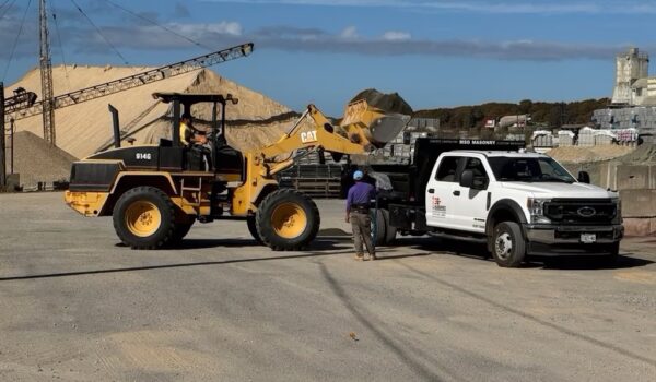 A construction loader depositing sand onto a flatbed truck labeled "MSO Masonry" at an industrial yard with sand piles and conveyor belts in the background.